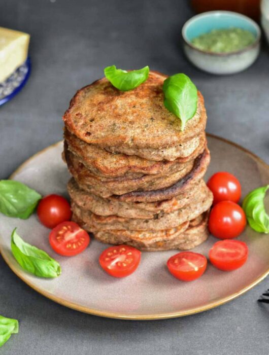 A stack of buckwheat pancakes on a brown plate with cherry tomatoes and basil leaves.