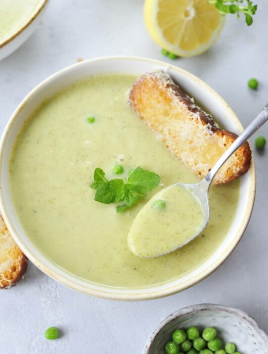 white plate with zucchini soup, spoon, mint leaves and cheese toast in the middle of the soup