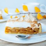 a piece of apple tart with meringue topping on a white plate, leaves in the background