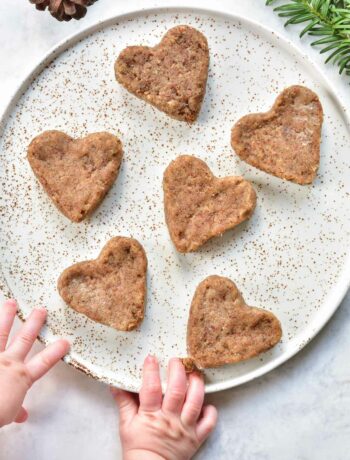 heart-shaped soft gingerbread cookies for babies on a white plate, baby hands reaching for cookie