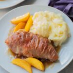 Prosciutto-wrapped chicken with peaches and mashed potatoes on a blue plate.