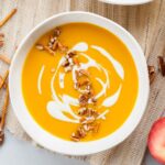 White bowl with butternut squash and apple soup topped with yogurt, pretzel, and pecans.