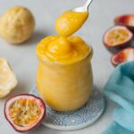 Passion fruit curd is being spooned into a jar. Passion fruits and a blue kitchen cloth in the background.