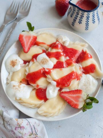 Sweet cheese pierogi on a white plate topped with strawberry sauce and whipped cream.