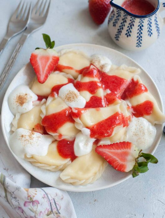 Sweet cheese pierogi on a white plate topped with strawberry sauce and whipped cream.