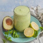 Avocado cilantro lime dressing in a jar on a green plate surrounded with avocado, limes and cilantro.