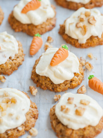 Carrot cake cookies with cream cheese frosting on a piece of parchment paper topped with marzipan carrots and nuts.