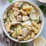 Chicken zucchini pasta in a white bowl. Lemons, parsley, and a kitchen cloth in the background.