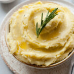 Cheddar mashed potatoes in a white bowl topped with a rosemary sprig.