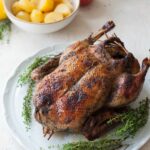 Roast duck on a white plate. Apples and potatoes in the background.