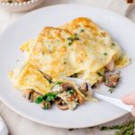 Two mushroom filled crepes in a beige plate are being cut with a fork.